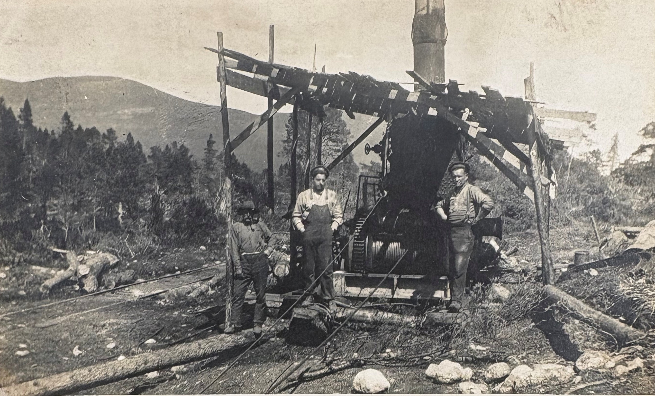 A black-and-white historical photograph showing three individuals standing outdoors beside a large piece of industrial machinery, likely a steam-powered winch or engine, housed under a makeshift wooden shelter with a slanted roof. The machinery has a prominent cylindrical component and a tall chimney extending upward. The ground is rough and scattered with logs, ropes, and stones, suggesting a logging or forestry operation. In the background, there are trees and rolling hills, indicating a remote, mountainous location. The individuals are dressed in work clothes typical of the early 20th century.
