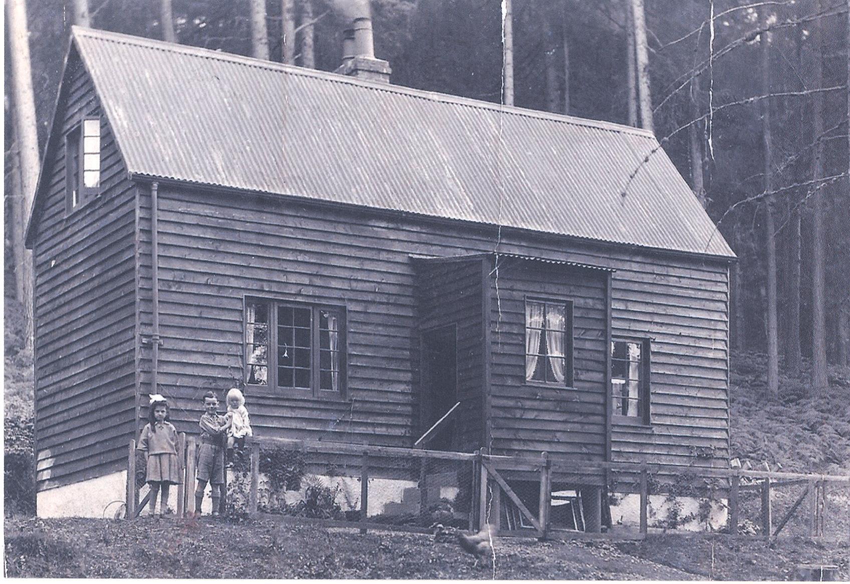 A black-and-white photograph of a small wooden house with a steep, corrugated metal roof and a chimney. The house has two visible windows with curtains and a central doorway, accessed by a short set of steps. A simple wooden fence runs along the front of the house. In front of the fence stand three children, dressed in early 20th-century clothing. The background shows tall trees, suggesting the house is located in a forested or rural area.