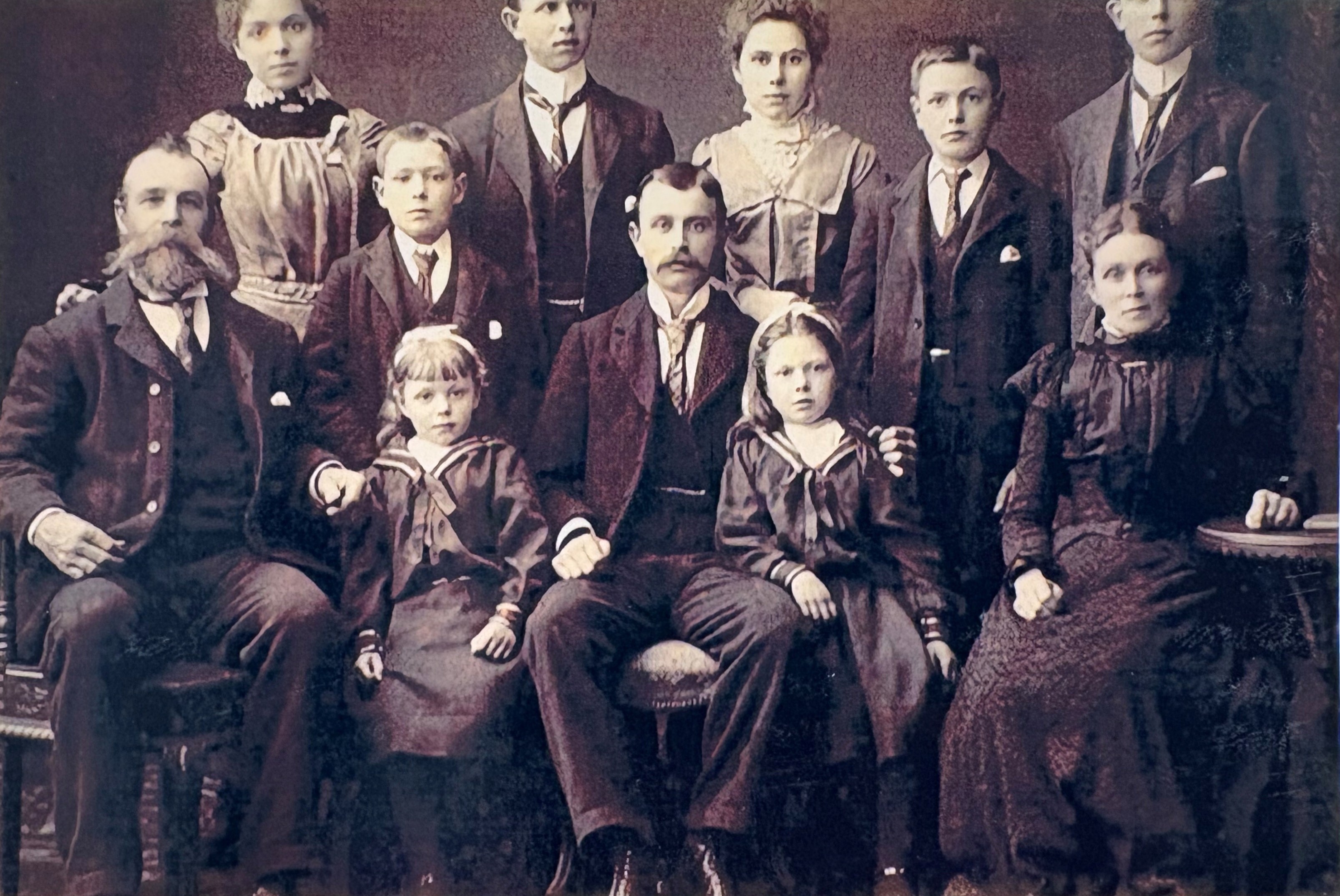 A sepia-toned vintage family portrait featuring a group of ten individuals arranged in two rows. The front row includes four seated figures, with two children in sailor-style outfits positioned in the center. The back row consists of six standing figures dressed in formal Victorian-era clothing, including high-collared dresses and dark suits with waistcoats. The overall setting suggests a studio photograph with a plain backdrop and ornate chairs.