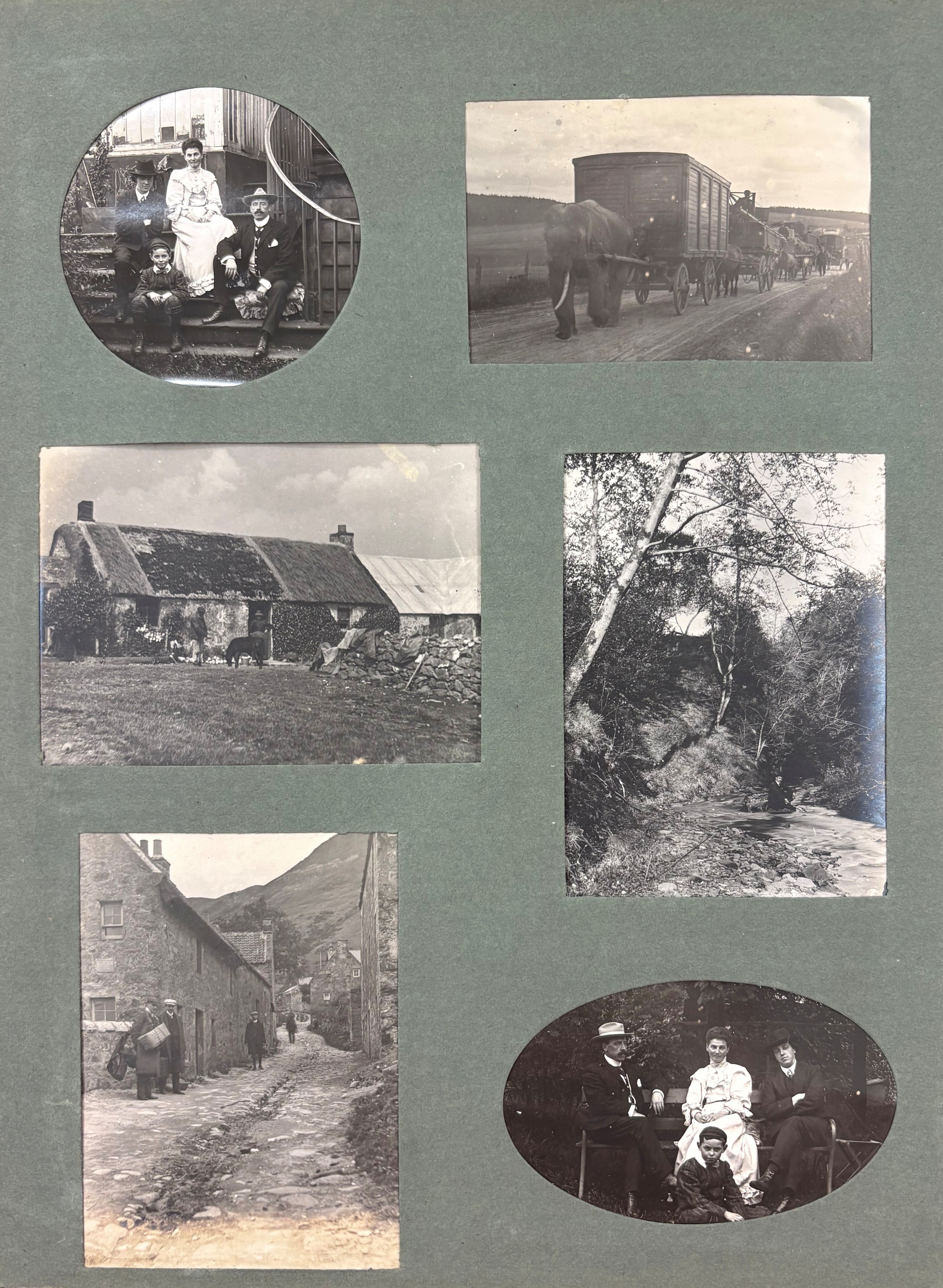 A scrapbook page featuring six black-and-white photographs arranged on a green background. The images include: a circular photo of three people seated on outdoor steps, dressed in formal clothing; a rectangular photo of a horse-drawn wagon train carrying large containers along a dirt road; a photo of a stone cottage with a thatched roof and a small flock of sheep in front; a photo of a narrow cobblestone street lined with stone buildings, with several people walking along it; a photo of a wooded stream with trees arching over the water; and an oval photo of three people seated outdoors, wearing formal attire and hats.
