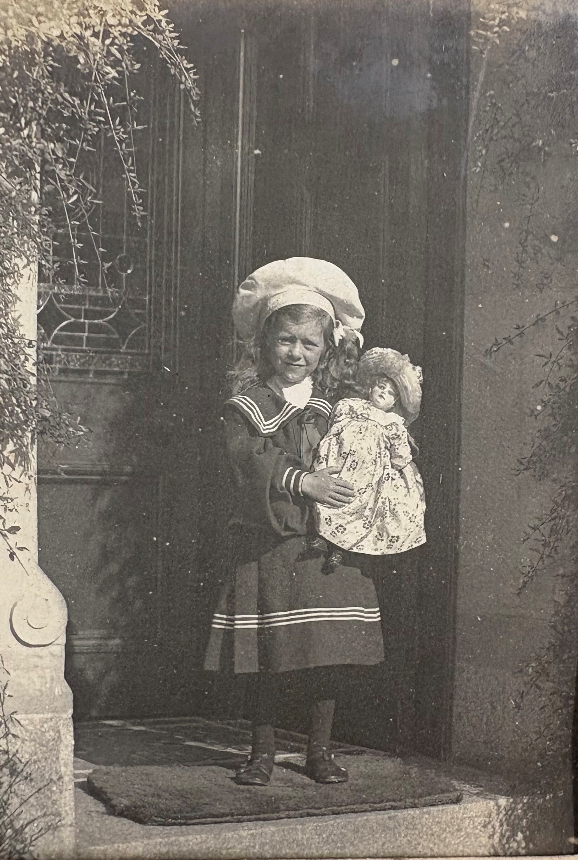 A vintage black-and-white photograph of a child standing in a doorway holding a large doll dressed in a patterned gown and bonnet. The child is wearing a sailor-style outfit with a wide-brimmed hat, dark stockings, and shoes. Decorative foliage frames the doorway on the left side, and part of a carved stone column is visible.