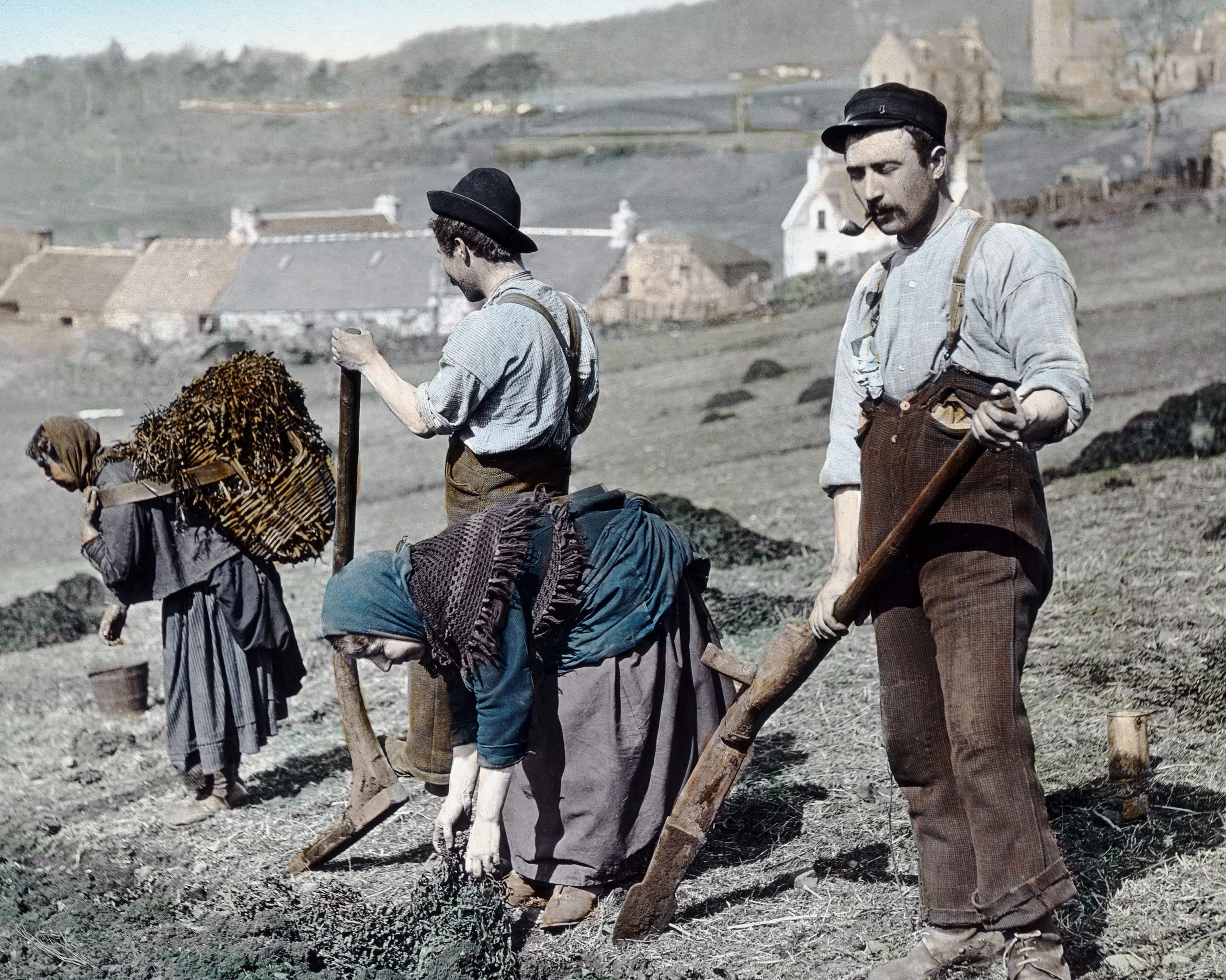 Colourised photograph showing a group of agricultural workers planting potatoes on sloping farmland. Three individuals in the foreground are using spades to dig and plant, wearing traditional work clothes including long skirts, aprons, and hats. One person carries a large wicker basket filled with material on their back. In the background, there are stone cottages and farm buildings scattered across the hillside, with open fields and distant hills beyond.