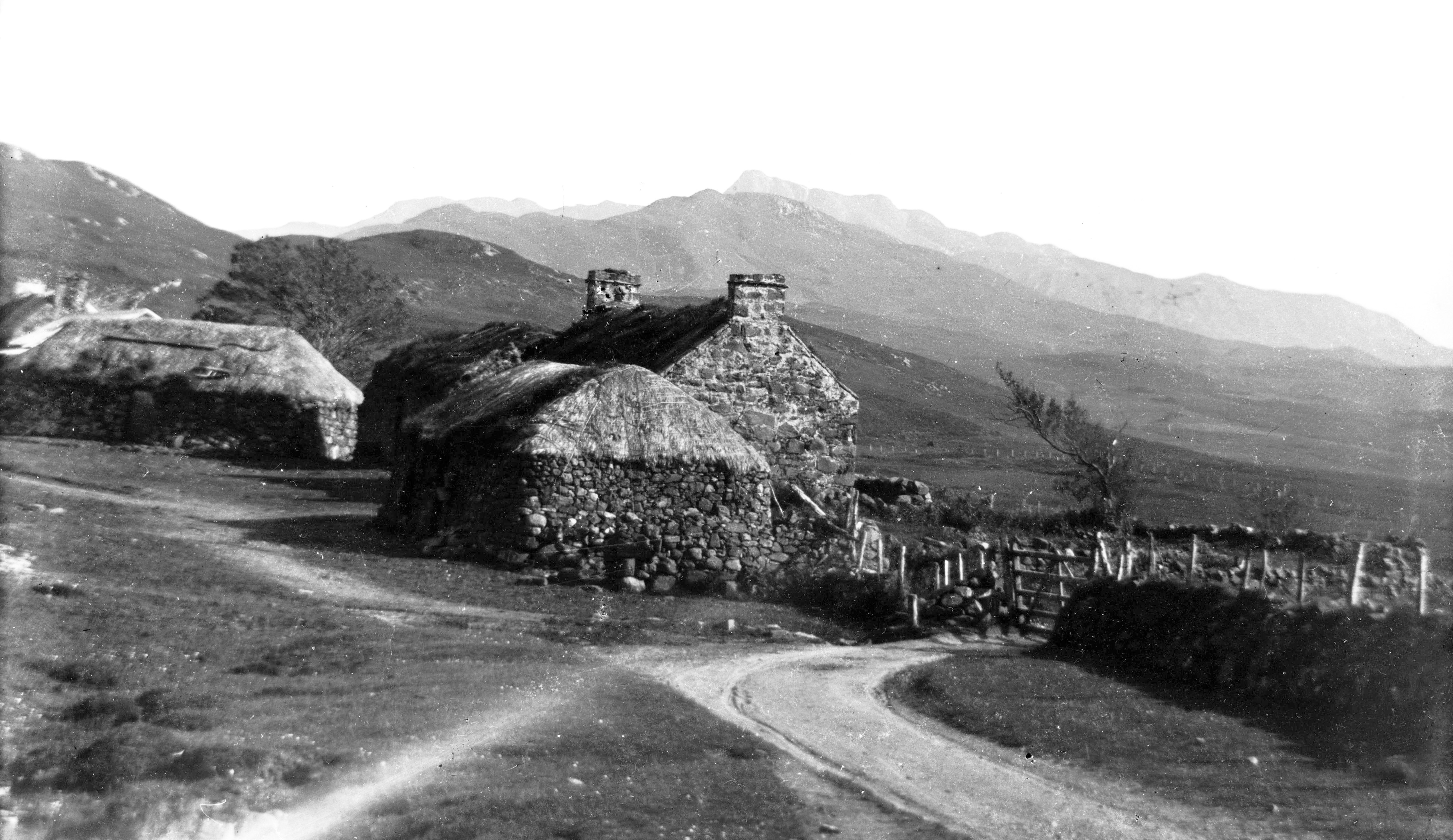 Black-and-white photograph of a small crofting township in a rural Highland landscape. The scene shows two traditional stone cottages with thick thatched roofs, surrounded by rough stone walls and a simple wooden gate. A winding dirt track curves through the foreground, leading toward the houses. In the background, rolling hills and rugged mountains rise under an open sky, creating a remote and picturesque setting.