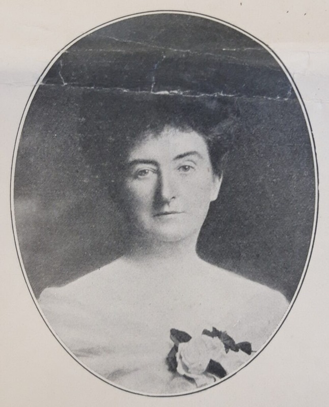 A black-and-white oval portrait photograph showing the upper body of a woman with fair skin and dark hair styled in a bun or chignon type style. She has a light-colored off the shoulder top on with a decorative floral accessory pinned on the left side of the chest. The background is dark and plain, and the image has visible signs of aging, such as creases across the top.