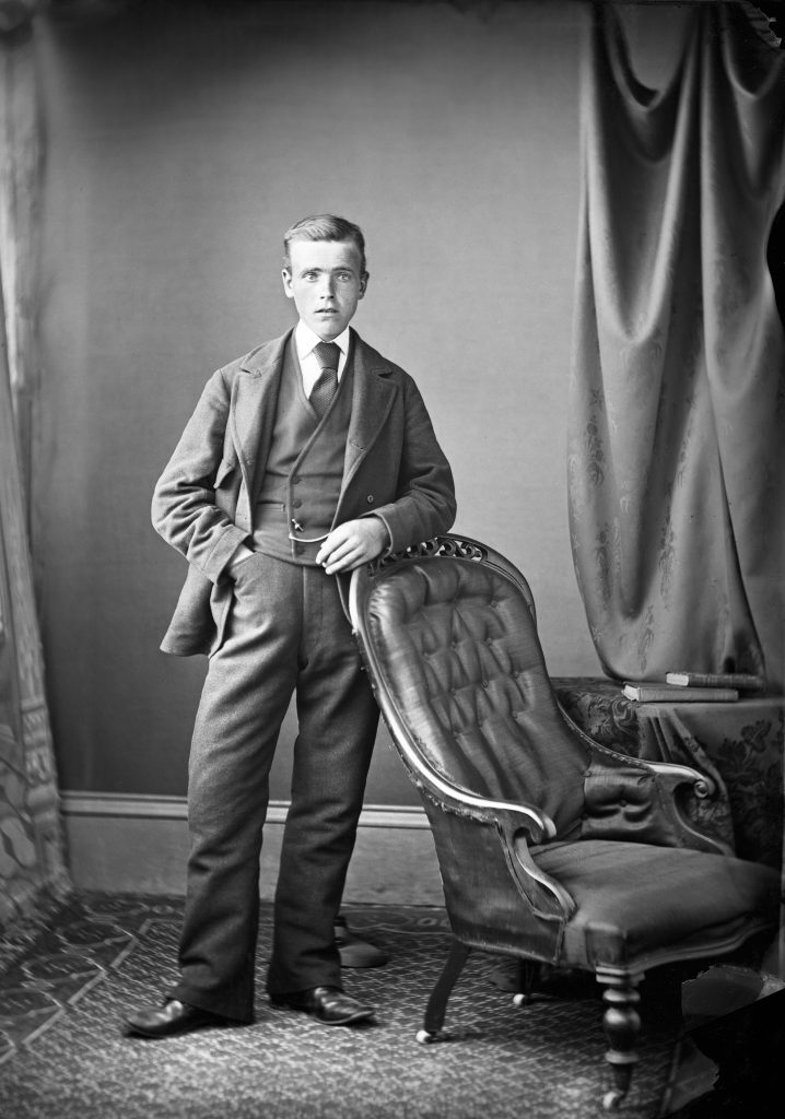 Portrait photograph of William Rae by Johnston photographers Wick. William is standing, in a three piece suit, with combed back hair, resting his arm upon the top of a leather chair.