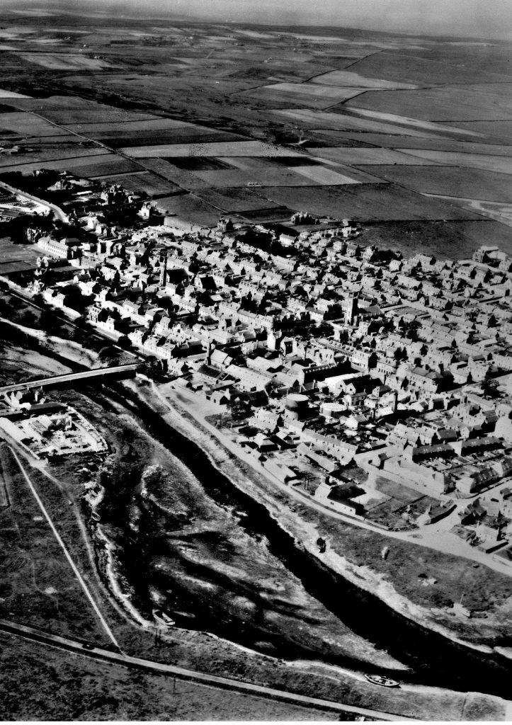 A black-and-white aerial photograph of a town with a river running through the foreground. A bridge spans the river, connecting two parts of the town. The town consists of densely packed buildings and streets, with larger structures interspersed among smaller houses. Beyond the town, expansive open fields and farmland stretch into the distance, creating a contrast between the urban area and rural landscape. The image captures the layout of the town, the river’s winding path, and the surrounding countryside.