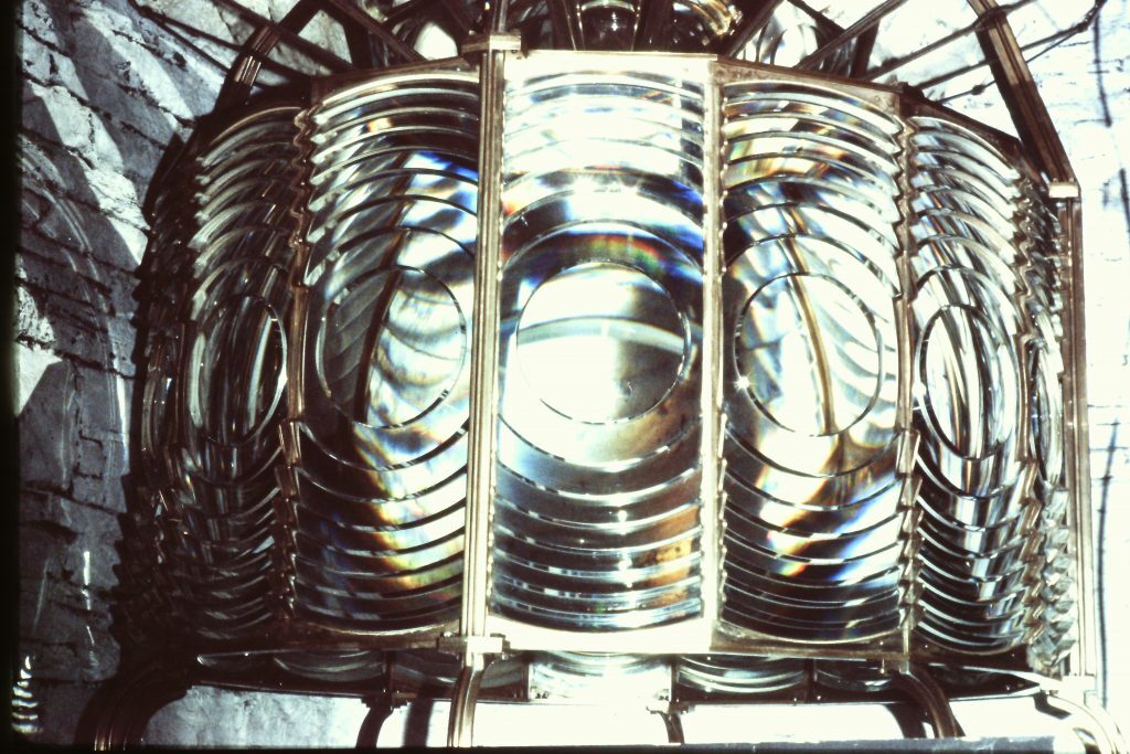 A large, intricate lens assembly. The lens consists of multiple concentric glass prisms arranged in a circular pattern, supported by a metal frame. Light reflects and refracts through the glass, creating rainbow-like colors. The background shows a stone wall with a rough texture.