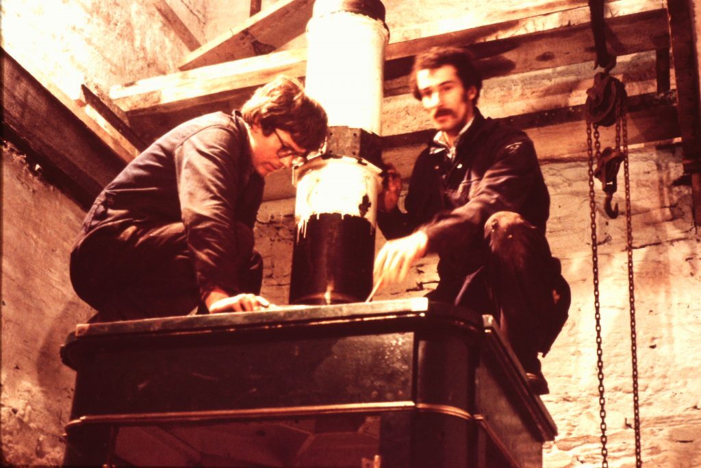Two individuals working on a large mechanical component inside a stone-walled structure. They are positioned on top of a dark metal base, handling a cylindrical part with white and black sections. Wooden beams and a hanging chain are visible in the background.