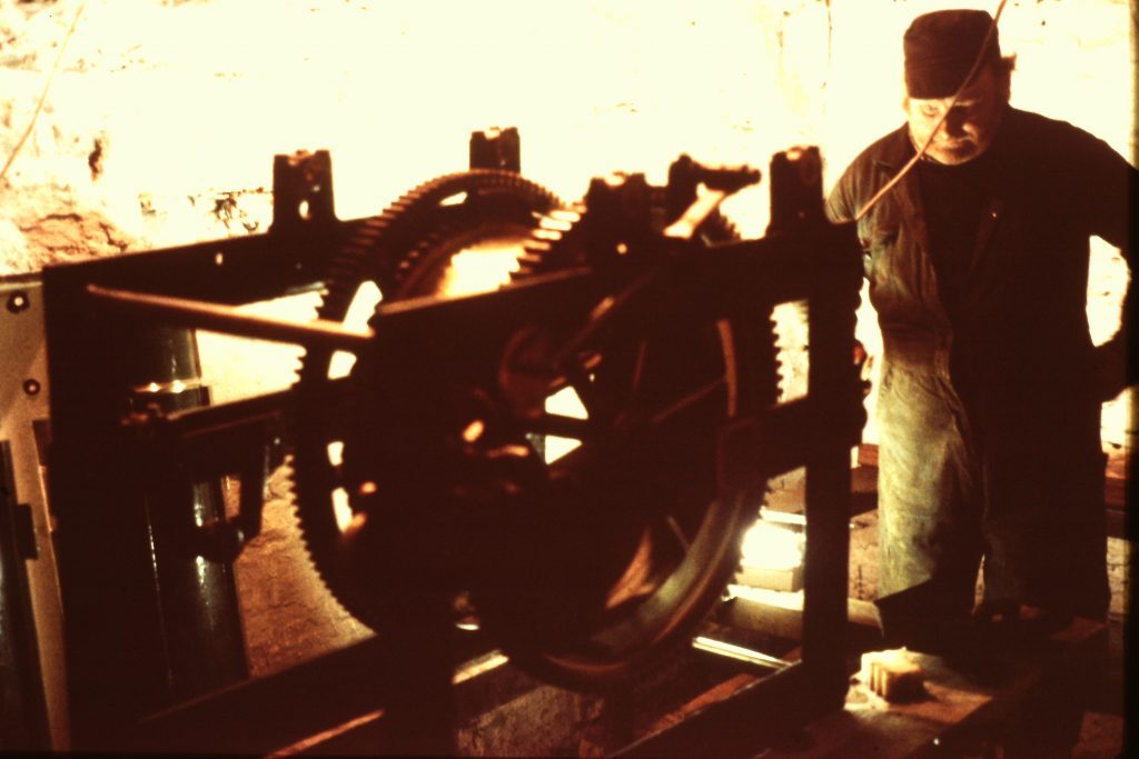 Person standing near a large mechanical assembly with visible gears and rods inside a stone-walled room. The machinery appears to be part of a clockwork or rotational mechanism, possibly related to lighthouse equipment. Bright light from behind casts the scene in silhouette.