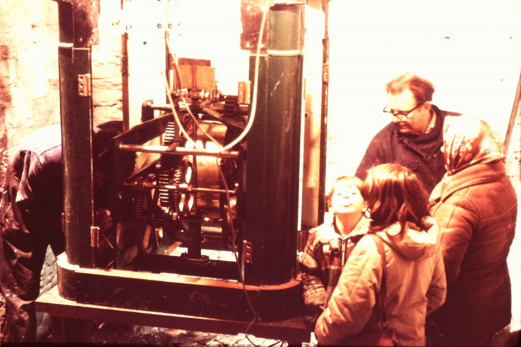 Group of people gathered around a large mechanical apparatus with visible gears and rods inside a green metal frame. The setting appears to be indoors with stone walls, and the machinery looks like part of a lighthouse mechanism or clockwork system.