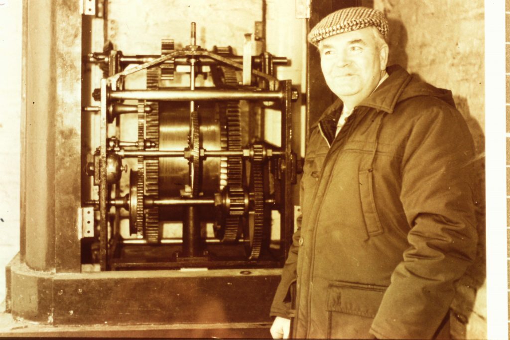 Large mechanical apparatus with multiple interlocking gears and cylindrical components housed within a sturdy metal frame, mounted on a base against a stone wall. A person wearing a heavy coat and patterned flat cap stands beside the machinery.