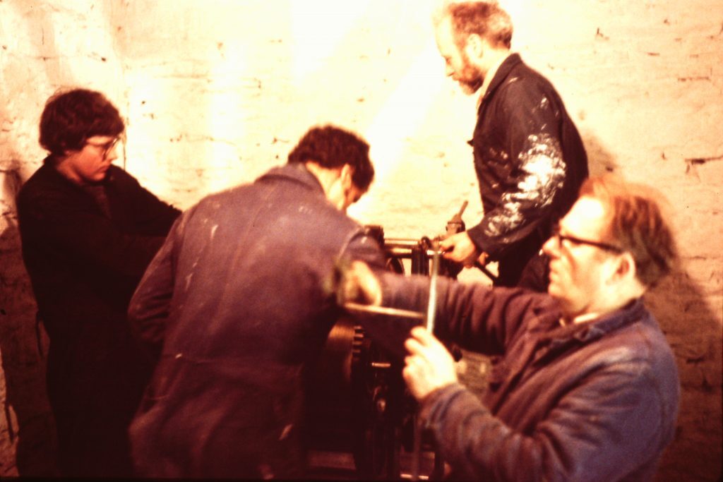 Four individuals working inside a stone-walled room on mechanical equipment. They are wearing work clothes and appear to be engaged in maintenance or assembly tasks. The equipment includes metal rods and a large cylindrical component.
