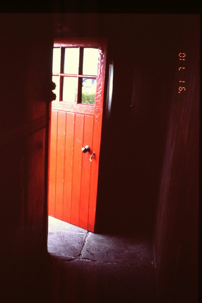 View from inside a dimly lit room looking toward an open red wooden door with a small window panel at the top. The door opens outward to reveal a glimpse of bright daylight and greenery outside. The floor inside is made of large stone slabs, and the surrounding walls are dark and wooden.
