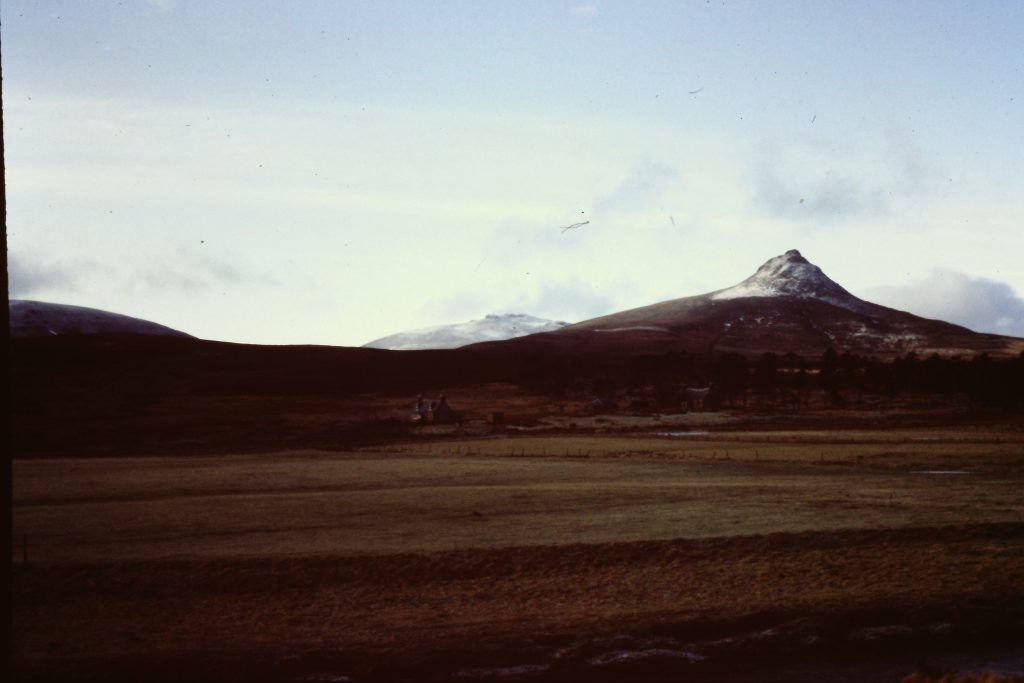 Landscape view of a rural area with open grassy fields in the foreground and a range of hills in the background. One prominent peak, partially dusted with snow, rises sharply against a pale blue sky with scattered clouds. A few small buildings and a cluster of trees are visible near the base of the hills.