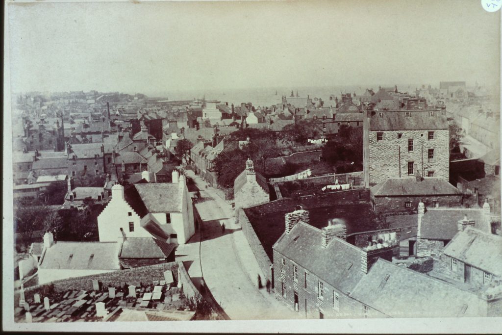 Historic sepia-toned photograph showing an elevated view of a coastal town with densely packed stone and slate-roofed buildings. A winding street runs through the center, flanked by houses and larger structures, including a prominent multi-story stone building on the right. In the foreground, a graveyard with rows of headstones is visible, and the background extends toward the sea with faint outlines of ships and harbor structures.