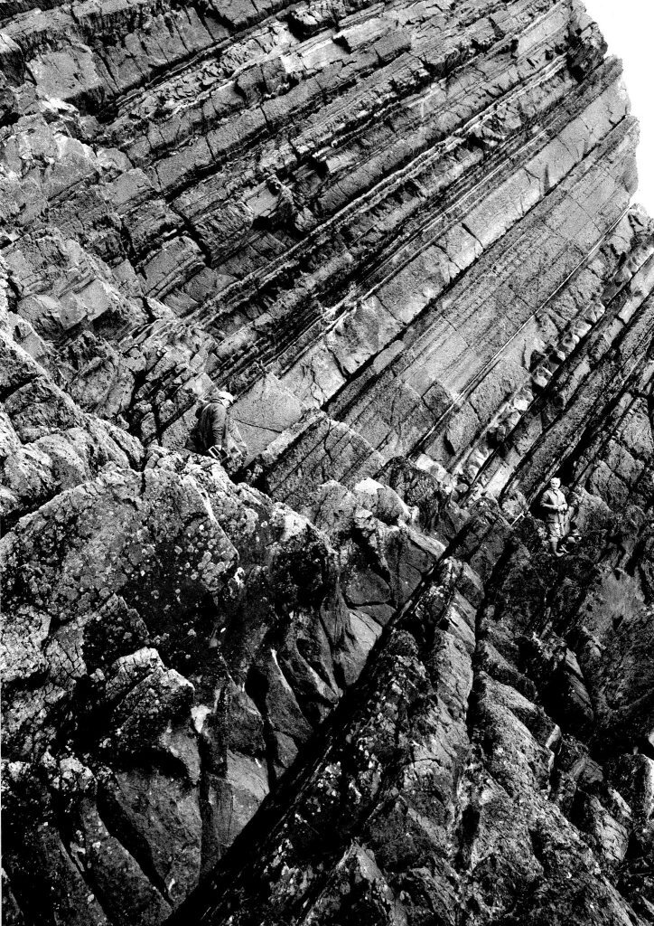 Black and white photograph of a steep, jagged rock face with prominent horizontal and diagonal striations. The textured surface shows layers of sedimentary rock, and a person wearing outdoor gear is climbing the formation using ropes, positioned toward the lower right section of the image.