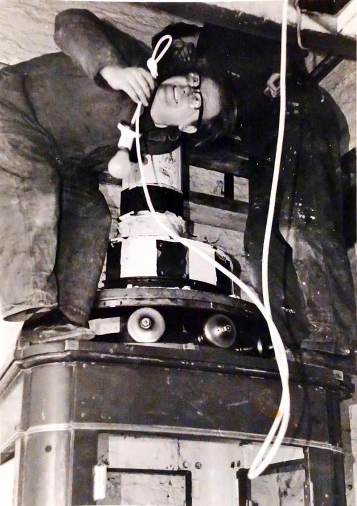A black-and-white photograph showing a person crouched inside a confined space, working on what appears to be a large mechanical or electrical device. The person is wearing glasses and practical work clothes, holding a light bulb attached to a cord. The device beneath them has a cylindrical section with alternating black and white stripes and several round components resembling bells or speakers. Wires and cables are visible, hanging loosely around the person and the equipment. The setting looks industrial or workshop-like, with rough walls and a sense of maintenance or construction work in progress.