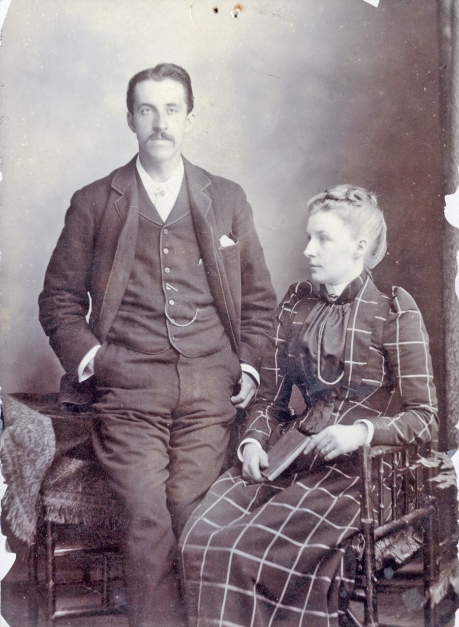 Black and white studio portrait of a man standing beside a seated woman. The man wears a three-piece suit with a pocket watch chain, and the woman wears a long checkered dress with puffed sleeves and holds a book in her lap. The photograph appears aged, with faded areas and worn edges.