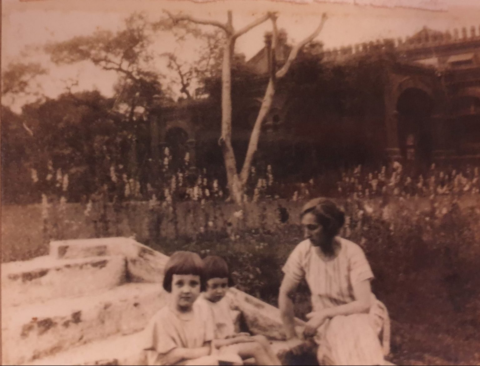 A sepia-toned photograph showing three people sitting outdoors on a stone structure in a garden. Two children are seated on the steps, and an adult is sitting beside them on the ground. Behind them is a large building with arched doorways and decorative architectural details, partially obscured by trees and tall flowering plants. A bare tree trunk stands prominently in the center of the image, adding contrast to the lush vegetation surrounding it. The overall scene suggests an early 20th-century setting in a landscaped garden.