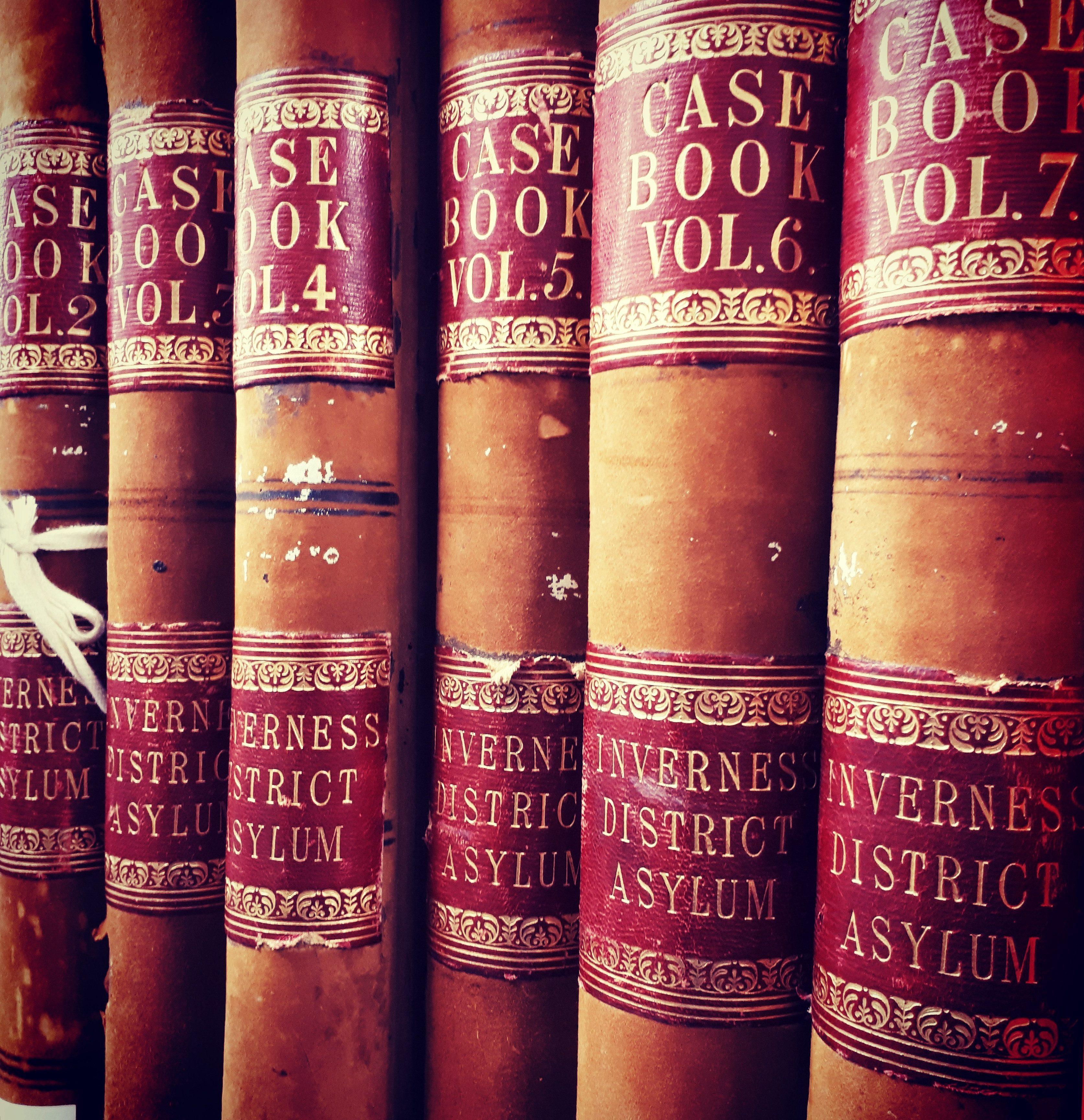 Close-up view of a row of large, leather-bound case books with red and gold labels on their spines. The titles read “CASE BOOK VOL.2,” “CASE BOOK VOL.4,” “CASE BOOK VOL.5,” “CASE BOOK VOL.6,” and “CASE BOOK VOL.7,” with “INVERNESS DISTRICT ASYLUM” printed below on each spine. The books show signs of age and wear.