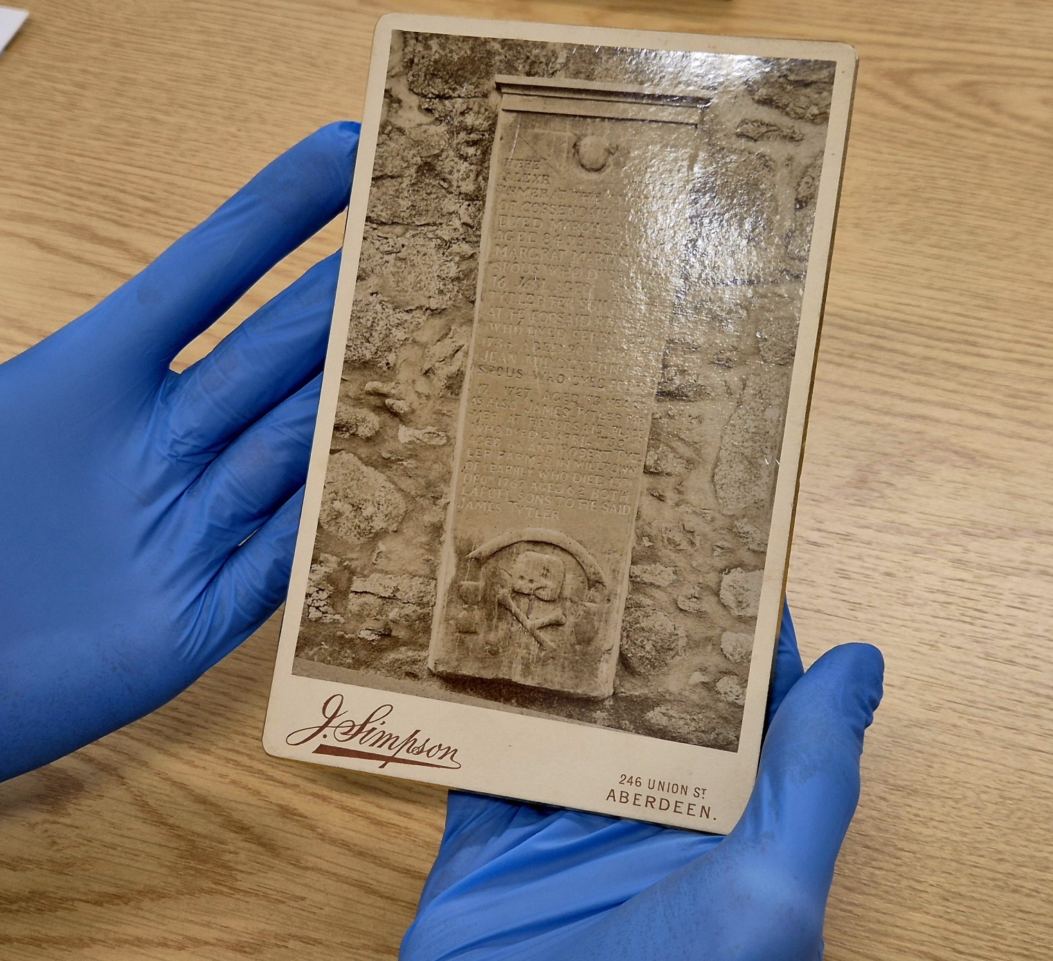 A person wearing blue nitrile gloves holds an old sepia-toned photographic card showing a stone memorial plaque set into a rough stone wall. The plaque is tall and rectangular with engraved text and a carved emblem near the bottom. The card bears the photographer’s imprint “J. Simpson, 246 Union St., Aberdeen.” The scene is set on a wooden table.