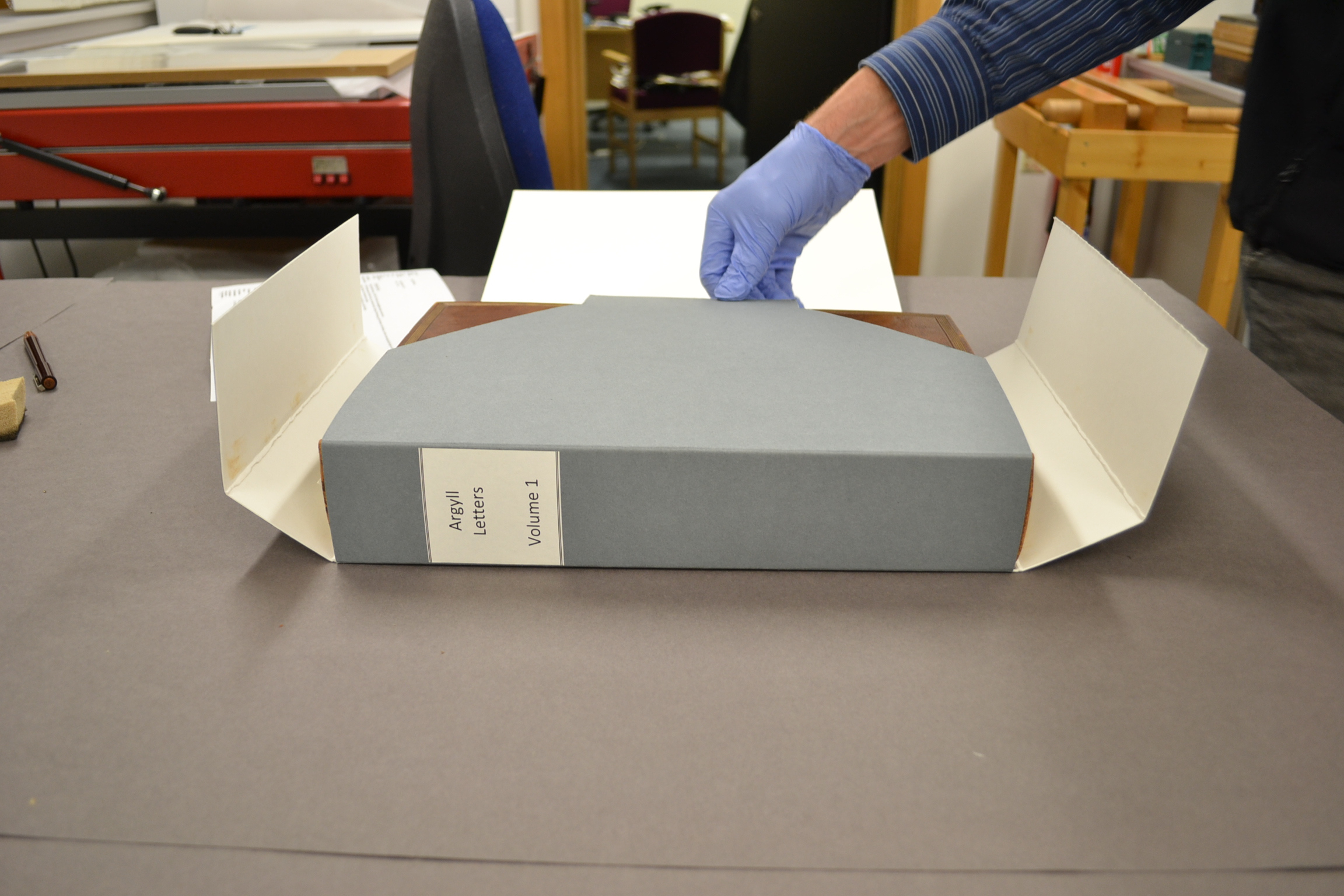A person wearing blue gloves is placing a grey archival storage box labeled “April Letters Volume 3” into a custom-made protective folder with flaps on a worktable. The setting includes conservation tools and materials in the background.