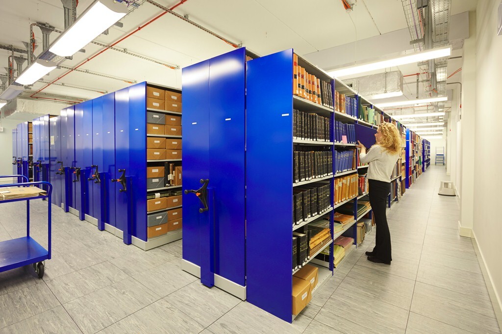 Modern archival storage room with high-density blue mobile shelving units, mechanical hand cranks, and organised shelves containing books, folders, and archival boxes. A person is replacing a book on the shelf.