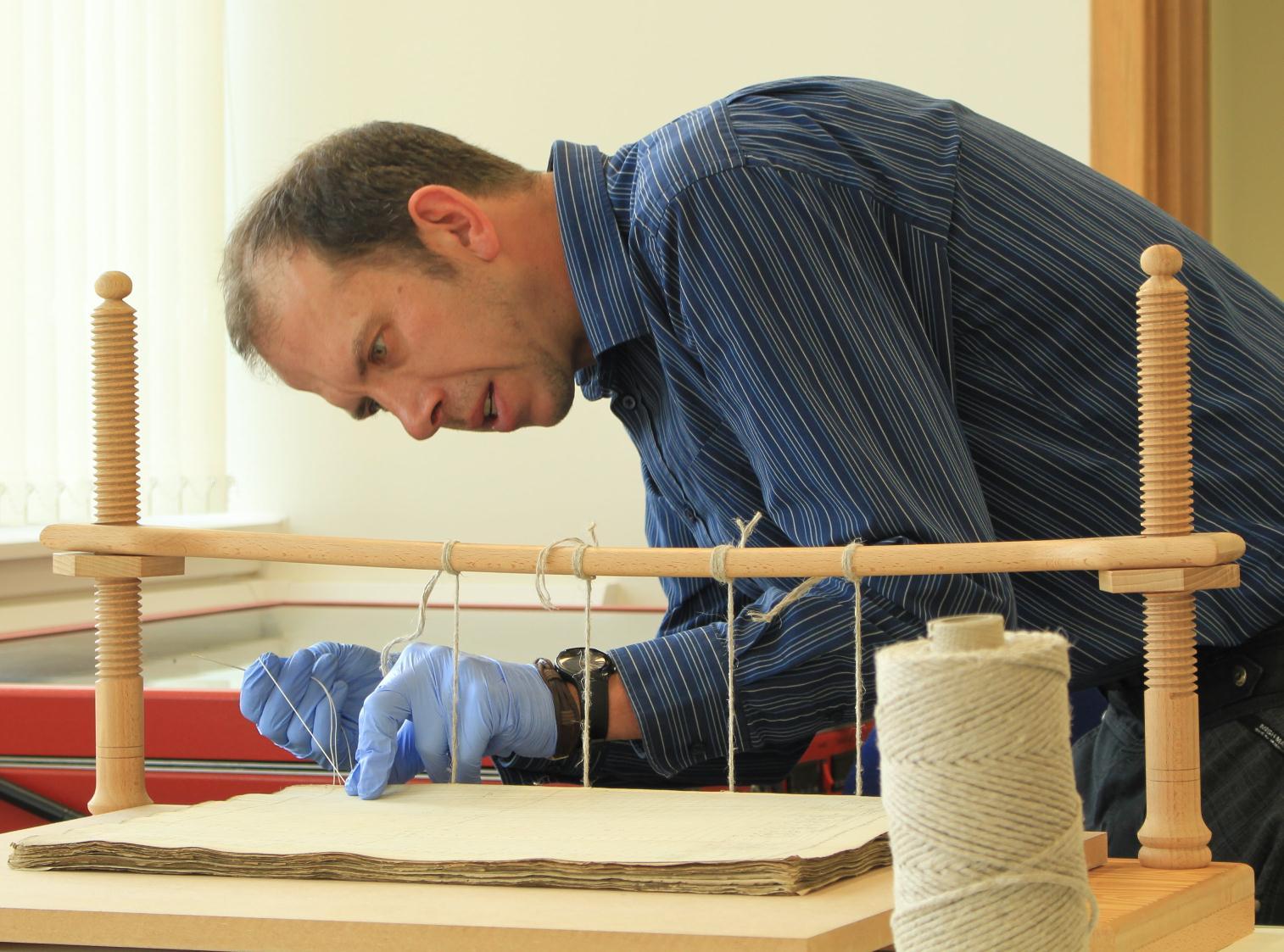 Person wearing blue gloves works on an open archival book held in a wooden book press, with a spool of thread on the table.