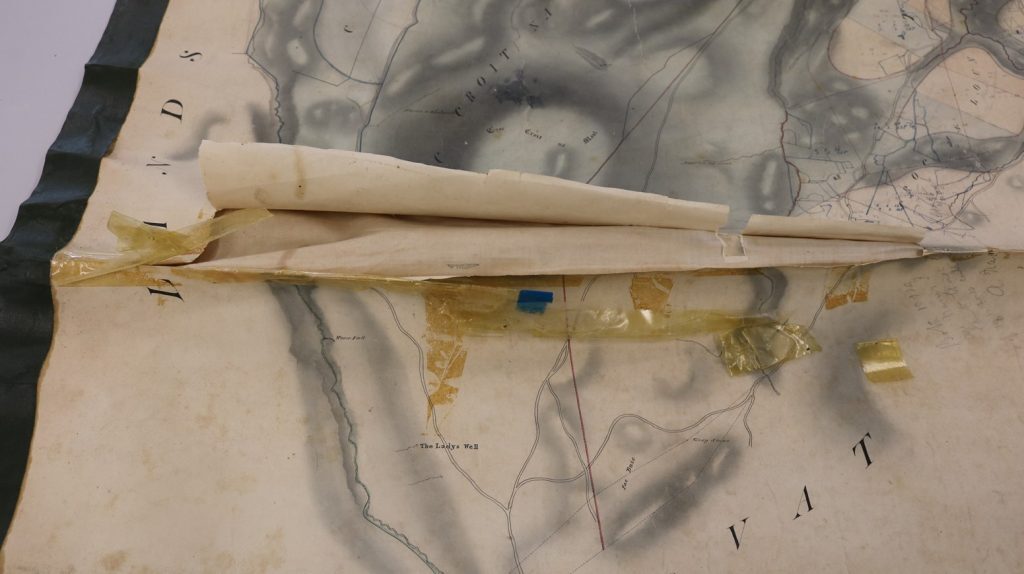 Close-up of a damaged historical map with a large folded section lifting from the surface. Several yellowed adhesive tape strips and a small blue patch are visible along the tear, indicating previous repairs. The map shows faded ink lines and shaded topography.