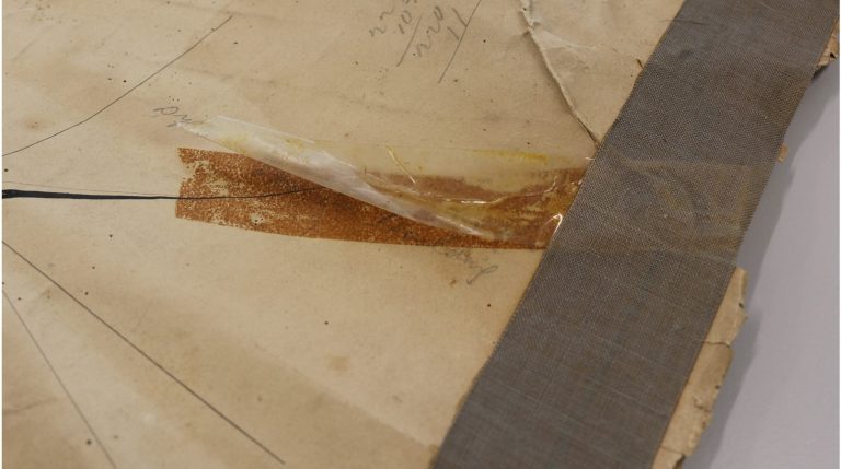 Close-up of a damaged map corner showing a wide grey cloth binding strip along the edge, a torn section with old adhesive tape partially lifted, and a large brown stain beneath the tape.