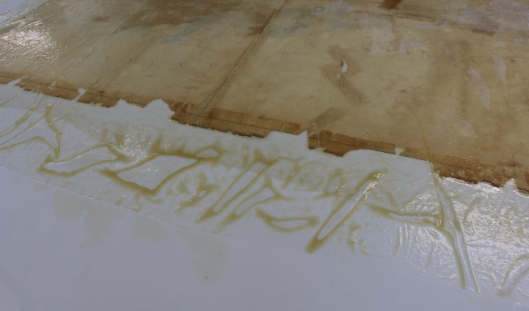 Close-up of a conservation worktable showing a historical map after wet cleaning. The surface is stained and discolored, with large areas of loosened dirt and adhesive residues visible on the white table around the edges of the map.