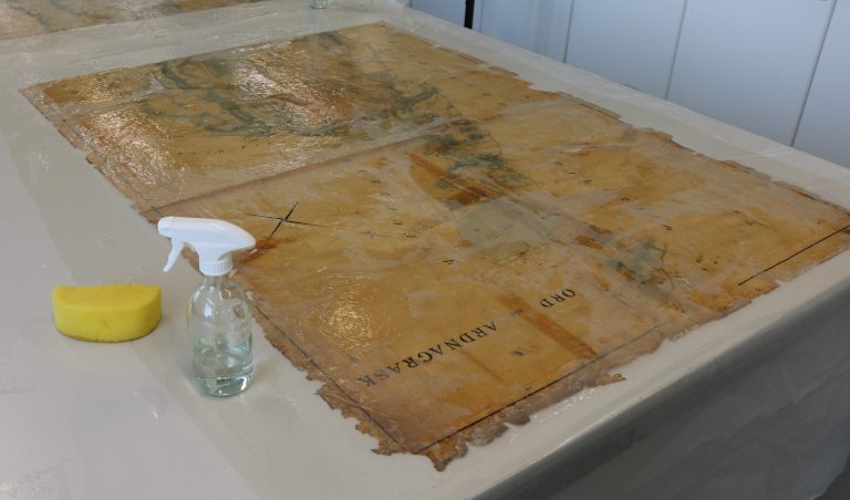 Historical map undergoing wet cleaning on a conservation worktable. The map appears darkened and stained, with softened surface layers from moisture. A yellow sponge and a spray bottle of water are placed nearby, indicating cleaning in progress.