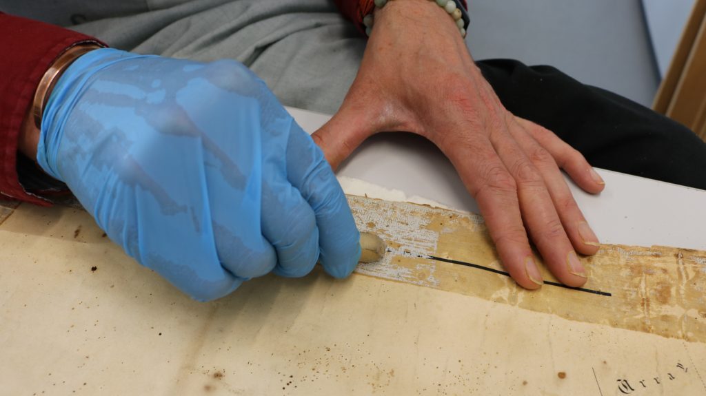 Close-up of a conservation process showing a person using a crepe eraser to remove old adhesive residue from a yellowed map. One hand wears a blue nitrile glove while the other steadies the paper. The map shows faded ink lines and scattered brown spots.