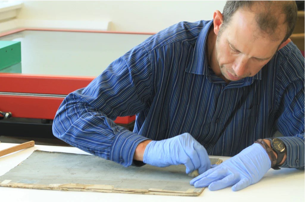 Person wearing blue gloves and a striped shirt carefully cleaning an old document on a work surface. A red archival storage unit is visible in the background.