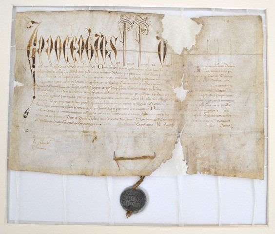 Restored historical parchment document with ornate Latin script at the top and smaller text below. The document shows repaired tears and areas of loss, mounted on a white support grid for stability. A dark wax seal hangs from the bottom edge.