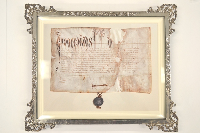 Framed digital copy of historical document featuring ornate Latin script at the top and smaller text below, with a dark wax seal attached at the bottom center. The frame is decorative with intricate silver detailing.