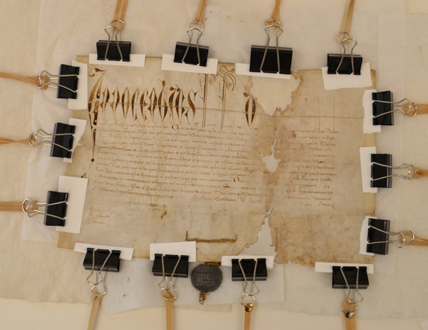 Historical parchment document with ornate Latin script at the top, secured on a work surface with multiple binder clips and wooden supports for drying after repair. The document shows patched areas and a dark wax seal attached at the bottom.
