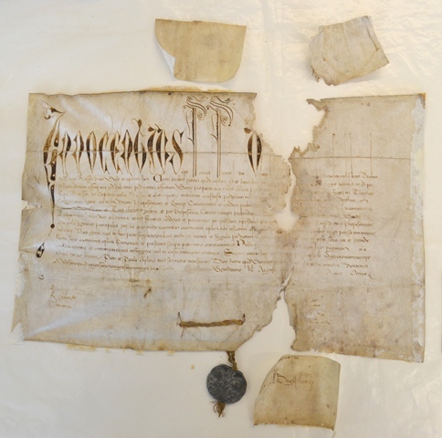 Historical parchment document with ornate Latin script at the top and smaller text below. The document shows heavy damage, including large tears, missing sections, and dark stains. Several old patch repairs have been removed and set to the side of the document, and a dark wax seal hangs from the bottom.