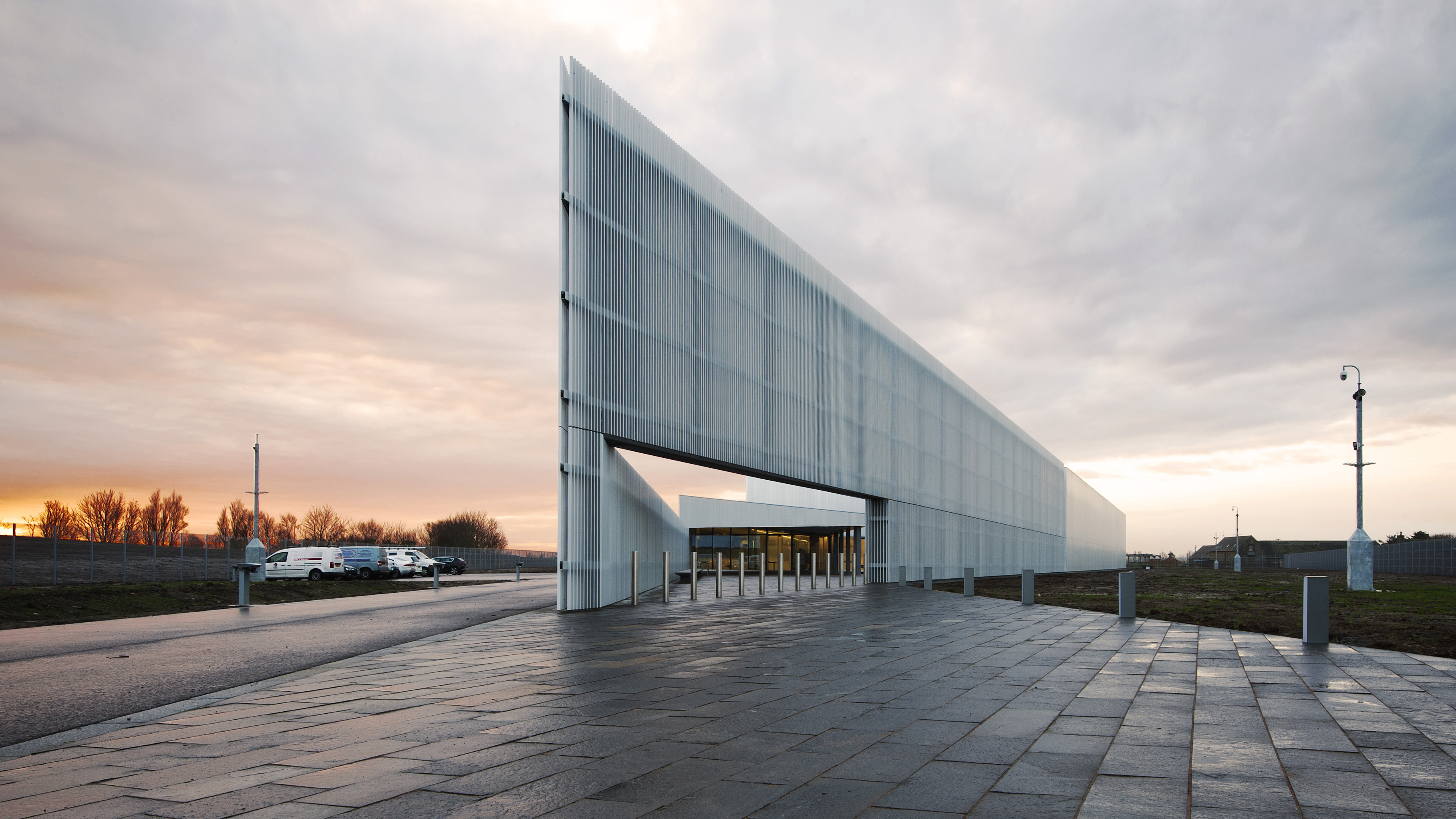 A modern building with a sharply angled facade shown against a dramatic cloudy sky.  The building is surrounded by a paved entrance