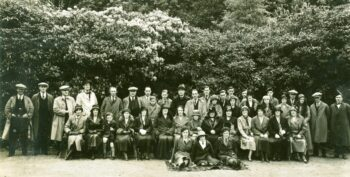 Black and white photograph showing a large group of people in formal dress and hats, sitting and standing in front of a backdrop of trees and bushes