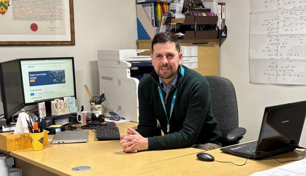 A man with dark hair sits at a desk with three laptops, a desktop monitor displaying the Scotland's People website, a printer, and various office supplies. Shelves with files and papers are in the background, and a large chart with handwritten notes is pinned to the wall.
