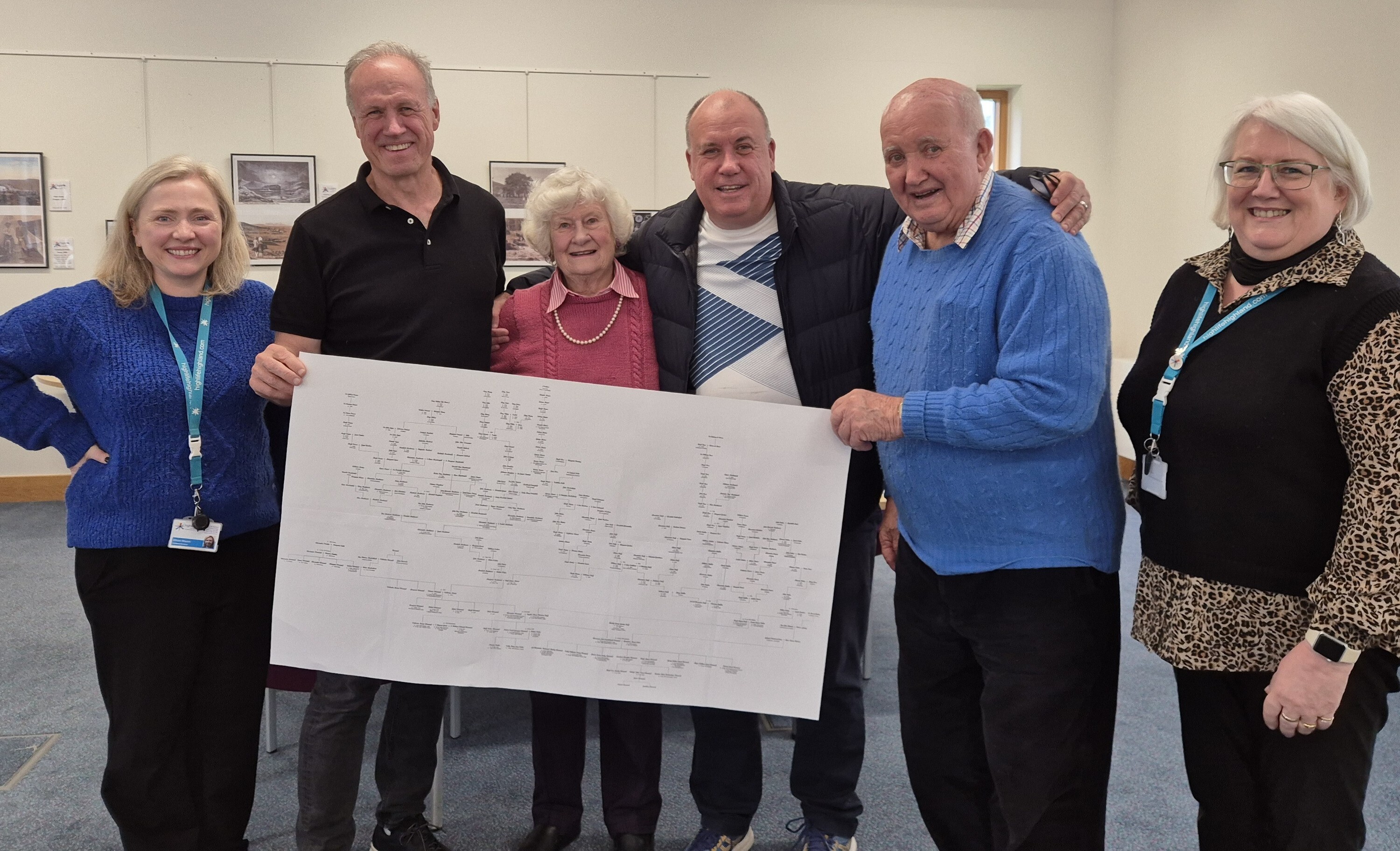 Four people standing indoors holding a large printed family tree chart. The chart contains numerous names arranged in a branching structure. The individuals are dressed in casual clothing, including sweaters and jackets, and are positioned in front of a light-colored wall with framed pictures hanging on it. The setting appears to be a library or archive search room with chairs visible in the background.