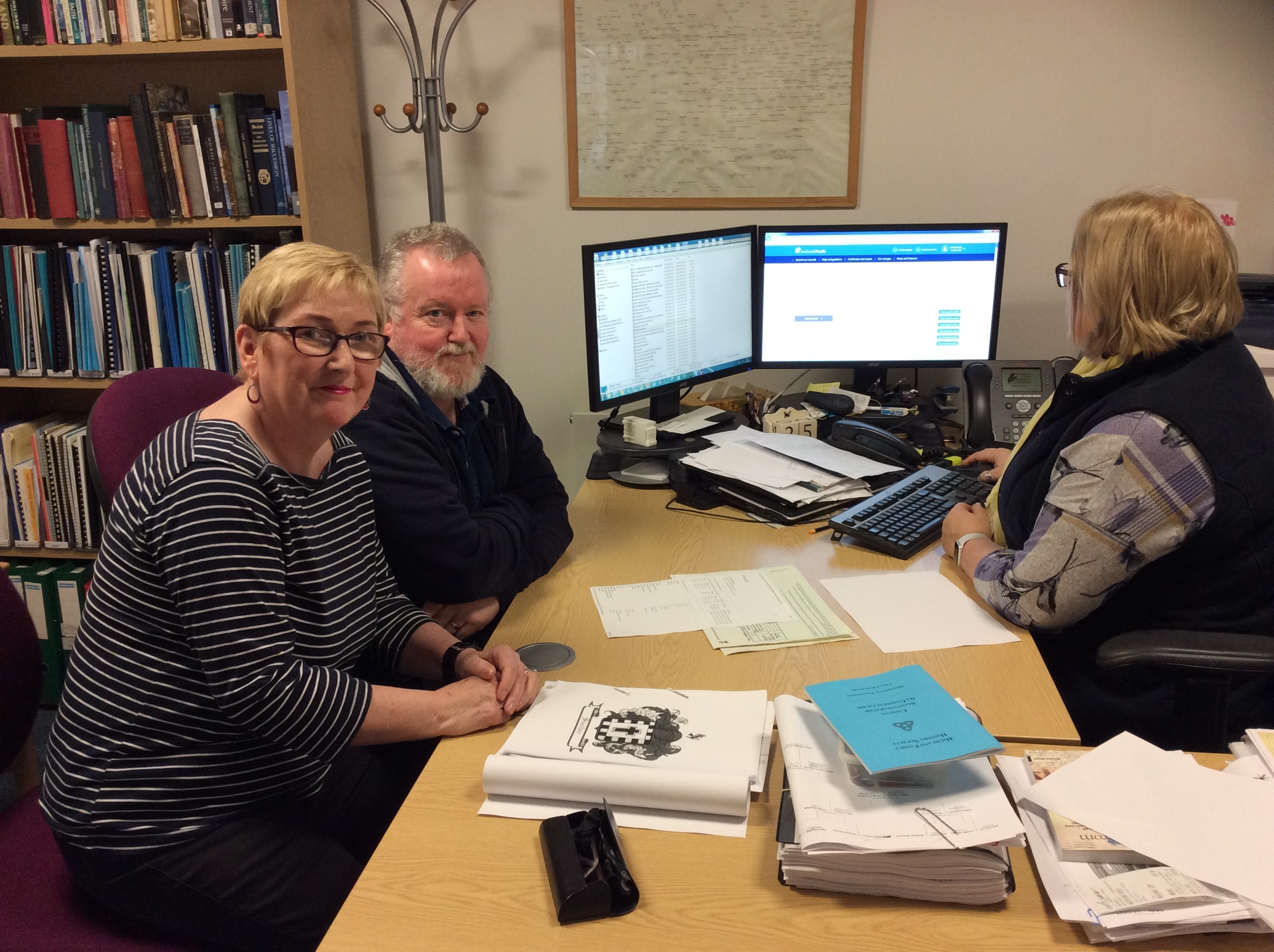 Two people sit at a desk across from an archivist who is working on a computer with dual monitors. The desk is covered with papers, folders, and printed documents, including a booklet with a crest on the cover. Behind the desk are bookshelves filled with books, and a large wall chart is partially visible.