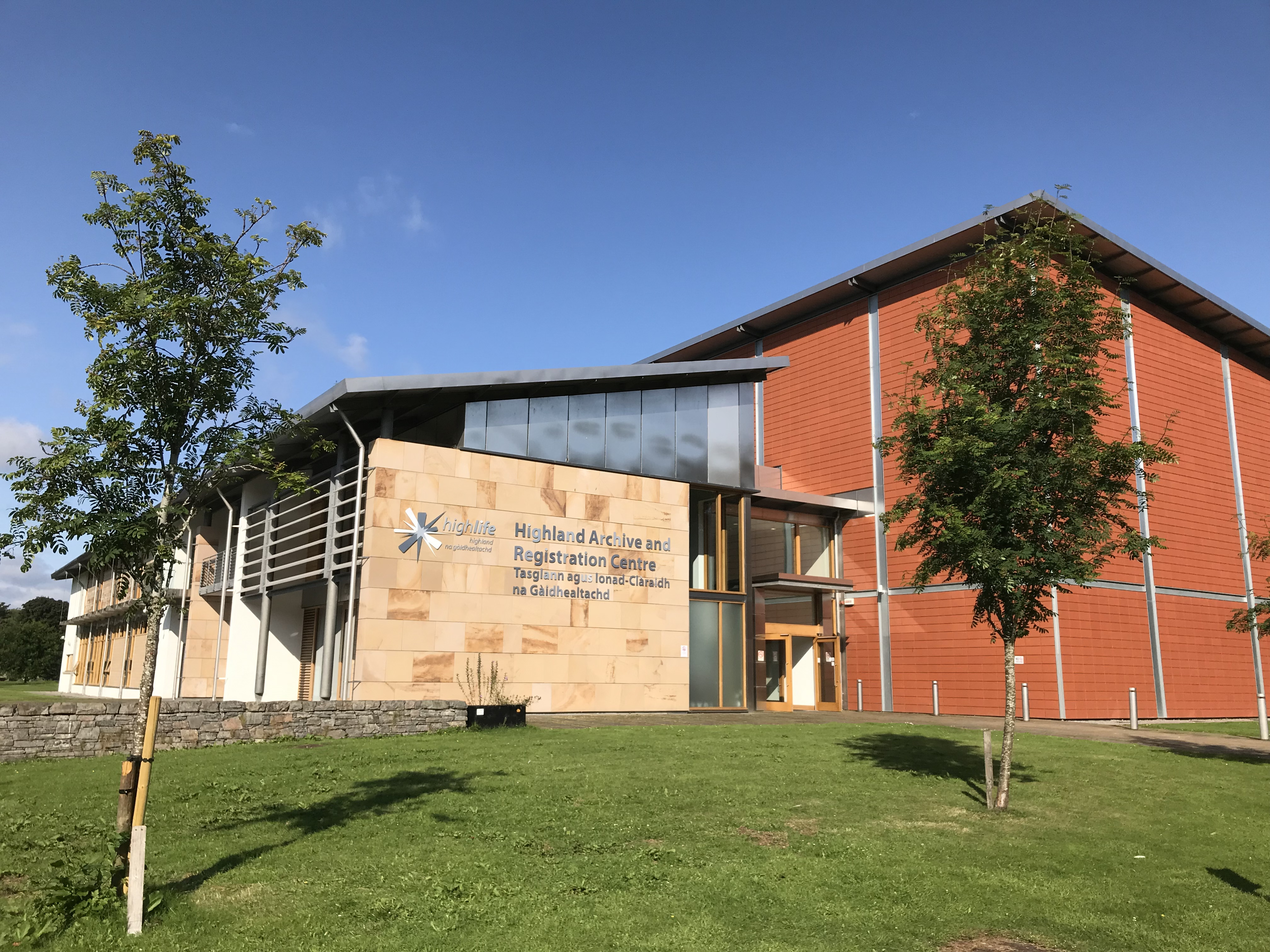 A modern building with light stone walls and large windows, labeled “Highland Archive and Registration Centre,” stands on a neatly maintained grassy lawn. Two small trees are in front of the building, and the sky above is bright and clear.