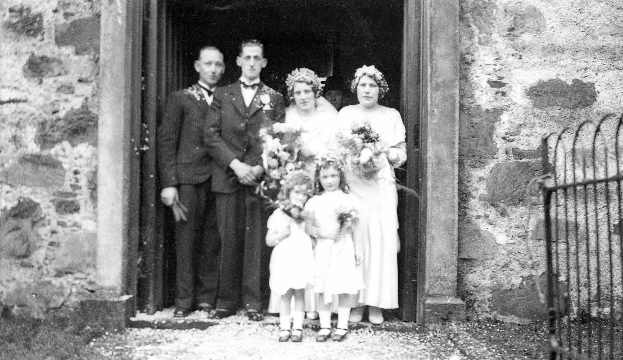The wedding of Eddie MacKenzie from Plockton to Lily Morrison from Totaig took place at Plockton Free Church. The wedding party are pictured standing at a door in a stone wall with metal railings to the right of the group. The best man, on the far left, is Angus Campbell, a shoemaker, and the bridesmaids are Katie and Alice MacKenzie, daughters of Duncan (Tailor) MacKenzie. The wedding party was held in Plockton Hall.