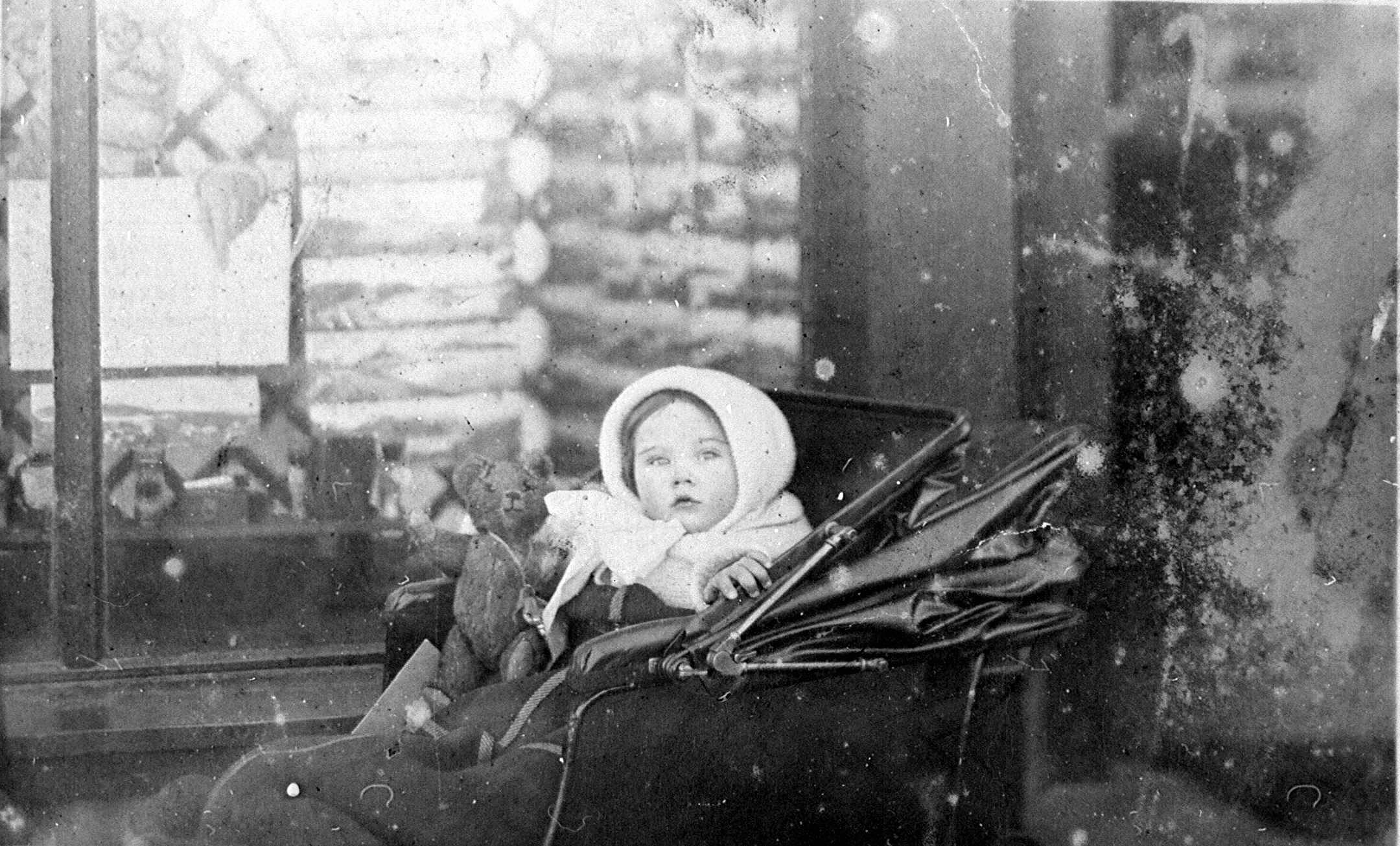 A vintage black‑and‑white photograph of a baby sitting in a pram indoors. The baby is bundled in warm clothing and holding a small soft toy. The pram has a folding hood and is positioned beside a window with patterned glass. The image has visible age marks and scratches. The baby is Mary, daughter of the photographer Duncan Macpherson and his wife Margaret. It was probably taken outside her father's pharmacy which he opened in Kyle of Lochalsh when he moved to the area in 1911.