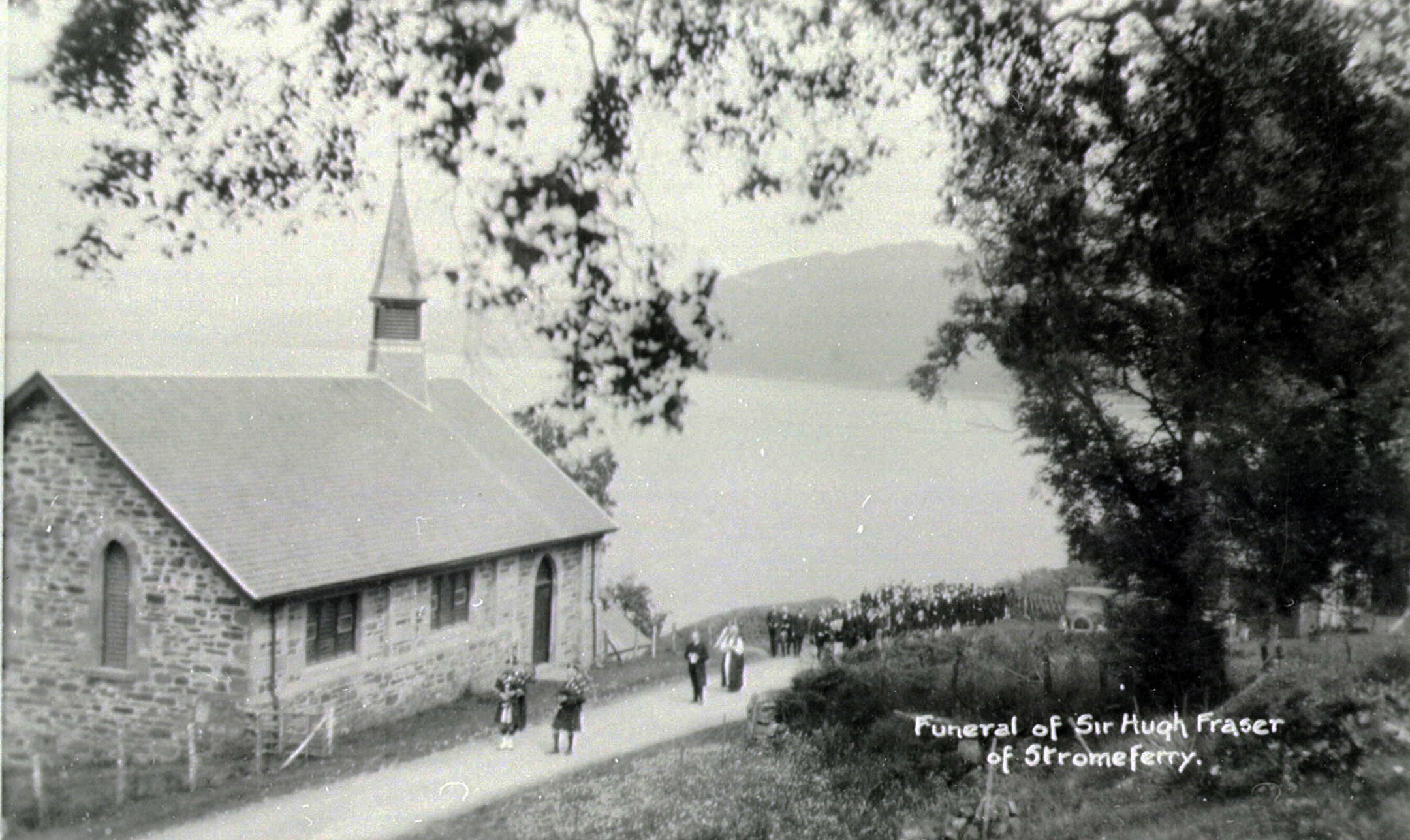 A black‑and‑white photograph showing a stone church with a steeple on a hillside road overlooking water and distant hills. A funeral procession walks along the road, with mourners gathered near the church and others following behind a hearse. Large trees frame the scene in the foreground. Text on the photograph reads “Funeral of Sir Hugh Fraser of Strome Ferry.”