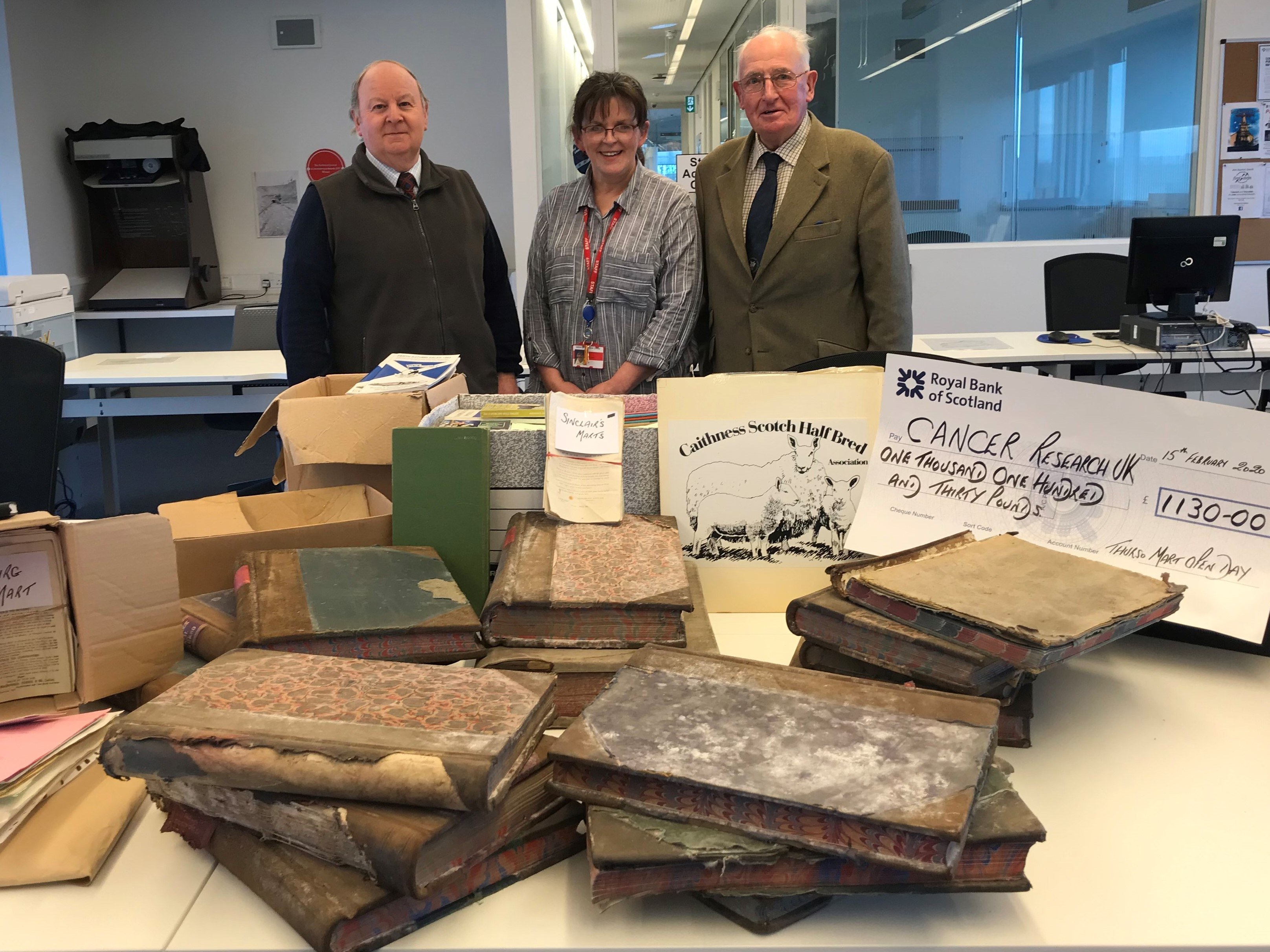 A woman and two men stand smiling behind a table covered with old books and archive material