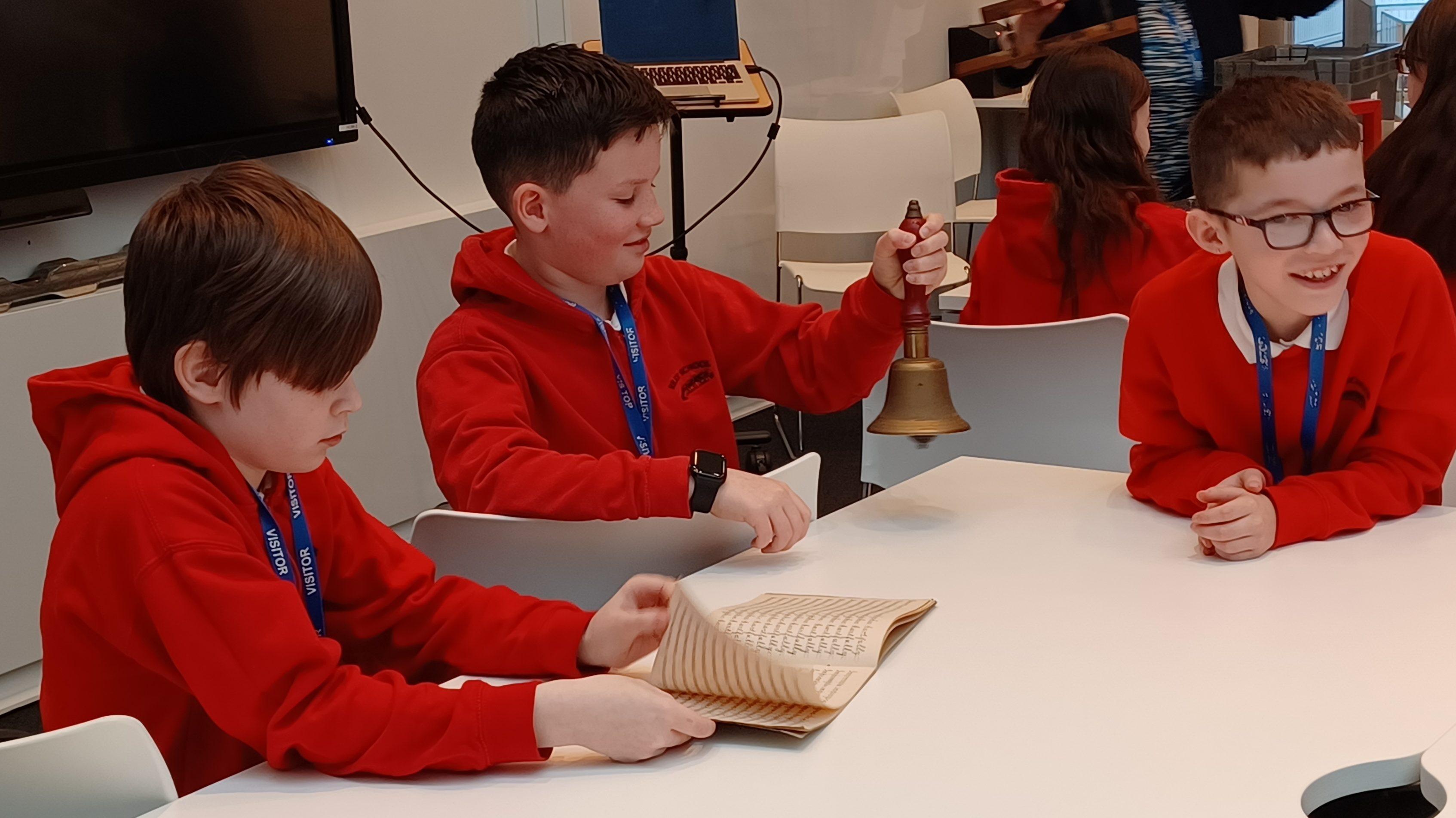 Three children in red school jumpers sitting at a white table looking at artefacts and documents