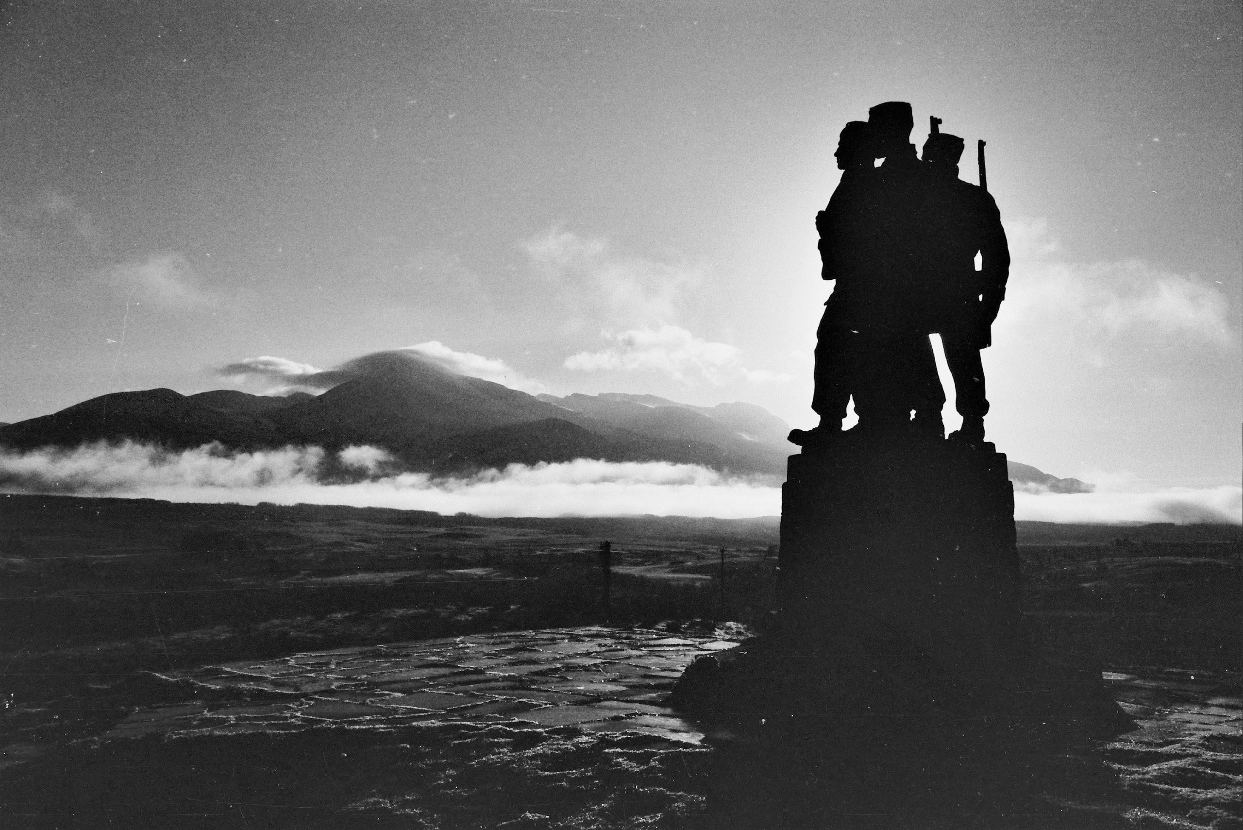 Black and white photograph showing the silhouette of a statues of three soldiers against a wide hilly landscape